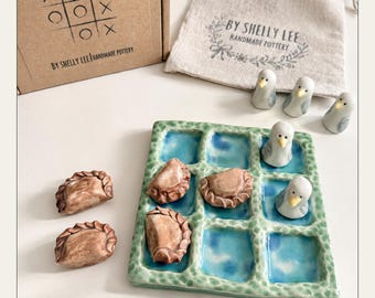 May include: A ceramic tic-tac-toe game set featuring a green grid board with blue squares, three brown clay dumpling-shaped pieces, and three white and gray bird-shaped pieces. Includes a cardboard box labeled TIC TAC TOE BY SHELLY LEE HANDMADE POTTERY and a linen drawstring bag with the same branding.