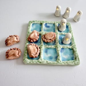 May include: A ceramic tic-tac-toe game set featuring a square board with a textured light green border and nine blue-glazed squares. The game pieces consist of five brown, crescent-shaped dumplings and four white, bird-shaped figurines.
