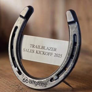 May include: A dark metallic horseshoe with a weathered finish, featuring the text 66 degrees engraved at the bottom. A rectangular metal plate is attached across the center, displaying the text TRAILBLAZER SALES KICKOFF 2025.