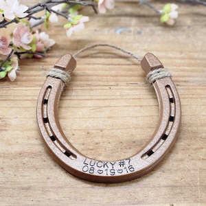 May include: A copper-colored horseshoe with a rope tied around it. The horseshoe is engraved with the text "LUCKY #7 08.01.2016".