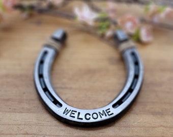 May include: A silver horseshoe with the word "WELCOME" engraved on it. The horseshoe is on a wooden surface.