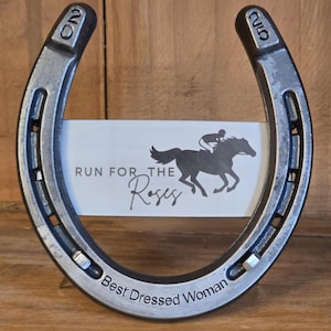 May include: A metal horseshoe with the numbers 20 and 25 stamped on the ends. The horseshoe frames a white rectangular card featuring a black silhouette of a horse and jockey with the text RUN FOR THE Roses and Best Dressed Woman.