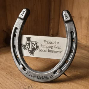 May include: A silver-toned metal horseshoe trophy featuring the numbers 24 and 25 stamped on the ends. A rectangular plaque is centered within the horseshoe, displaying the Texas A&M logo and the text Equestrian Jumping Seat Most Improved.