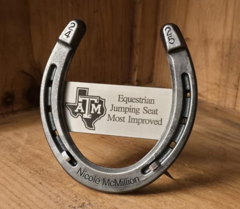 May include: A silver-toned metal horseshoe trophy featuring the numbers 24 and 25 stamped on the ends. A rectangular plaque is centered within the horseshoe, displaying the Texas A&M logo and the text Equestrian Jumping Seat Most Improved.