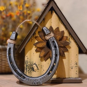Puede incluir: Una herradura plateada con las palabras "Bless Our Home" grabadas en ella. La herradura est&aacute; colgada de una casa para p&aacute;jaros con un dise&ntilde;o de girasol.