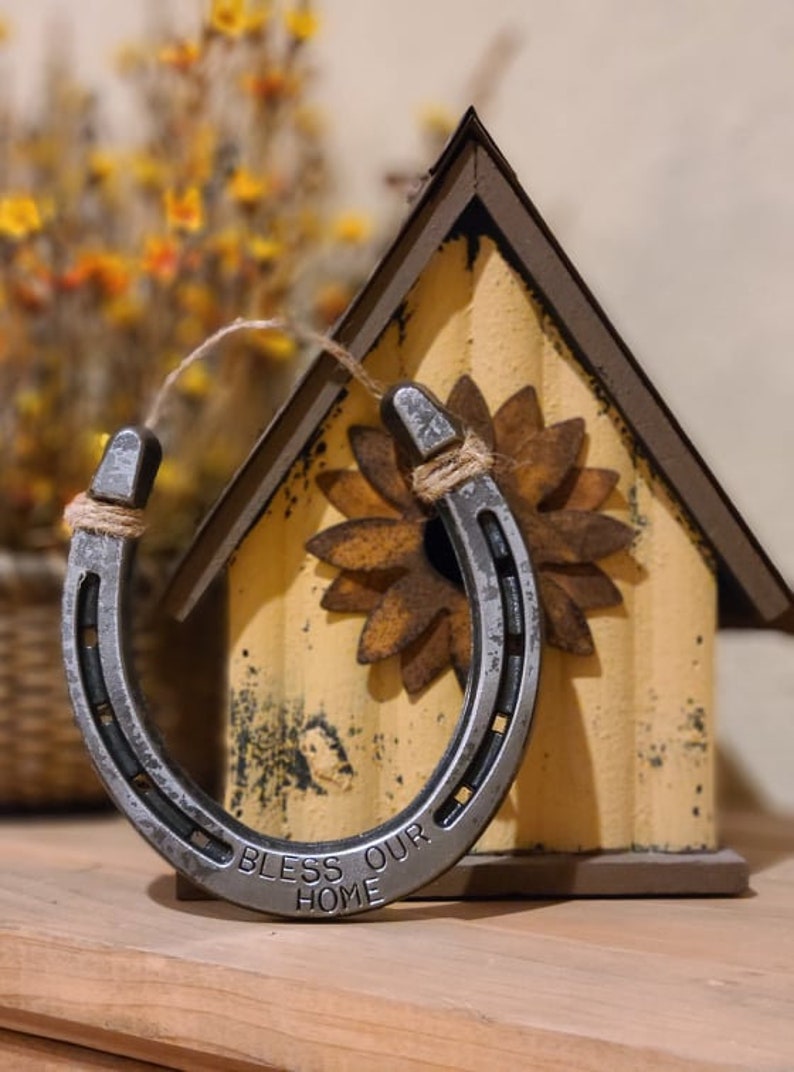 Puede incluir: Una herradura plateada con las palabras "Bless Our Home" grabadas en ella. La herradura est&aacute; colgada de una casa para p&aacute;jaros con un dise&ntilde;o de girasol.