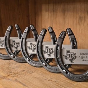 May include: A row of five metal horseshoe awards, each featuring a silver plate engraved with the Texas A&M logo and the text 'Equestrian Aggie Heart Award'. The horseshoes are stamped with '24' and '25' and personalized with names.