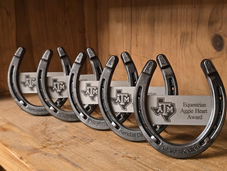 May include: A row of five metal horseshoe awards, each featuring a silver plate engraved with the Texas A&M logo and the text 'Equestrian Aggie Heart Award'. The horseshoes are stamped with '24' and '25' and personalized with names.