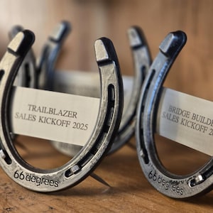 May include: Two silver horseshoes with the text "TRAILBLAZER SALES KICKOFF 2025" and "BRIDGE BUILDER SALES KICKOFF 2025" on white paper cards. The horseshoes are engraved with "66 degrees".