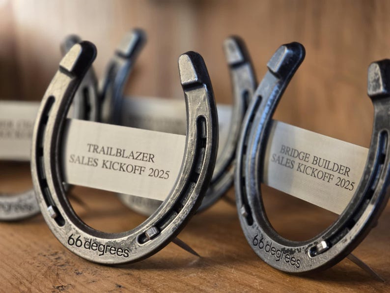 May include: Two silver horseshoes with the text "TRAILBLAZER SALES KICKOFF 2025" and "BRIDGE BUILDER SALES KICKOFF 2025" on white paper cards. The horseshoes are engraved with "66 degrees".