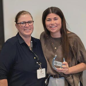 May include: Two individuals standing together, one wearing a dark navy collared shirt with a name tag and lanyard, the other in a brown short-sleeved top holding a silver horseshoe-shaped award with a small plaque featuring a logo and text.