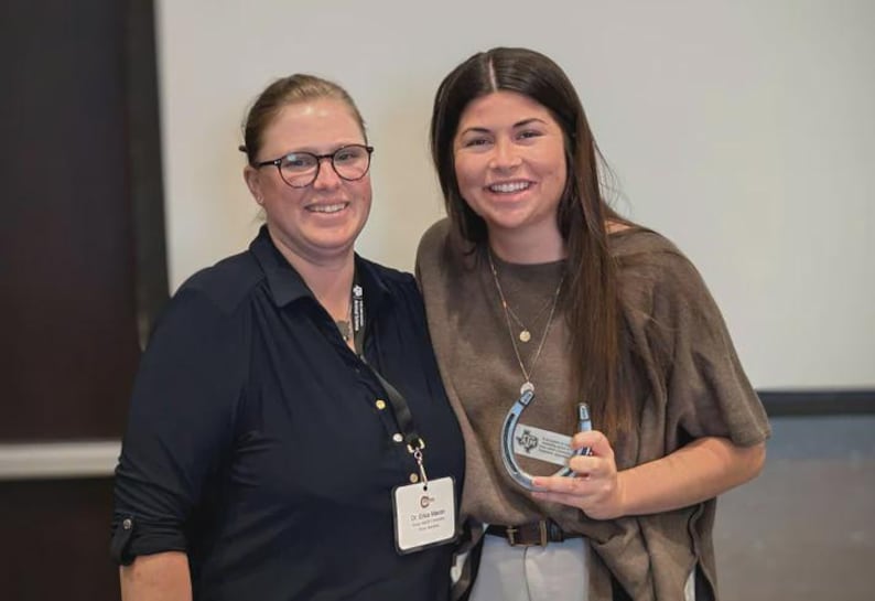 May include: Two individuals standing together, one wearing a dark navy collared shirt with a name tag and lanyard, the other in a brown short-sleeved top holding a silver horseshoe-shaped award with a small plaque featuring a logo and text.