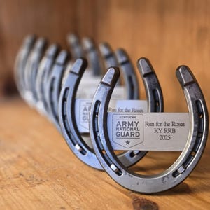 May include: A row of silver horseshoes with a white label attached to the front horseshoe. The label reads "Run for the Roses Kentucky Army National Guard KY RRB 2025".