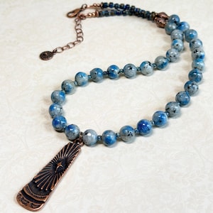 May include: A beaded necklace featuring blue and gray speckled stone beads. The necklace has a copper-colored pendant with a sunburst design. The clasp and chain are also copper-colored.