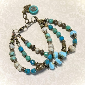 May include: A multi-strand bracelet featuring turquoise, blue, and off-white beads. The bracelet has antique brass-colored metal accents and a decorative clasp. The top strand has a turquoise charm with a floral design.