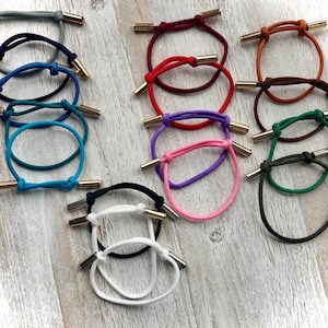 May include: A collection of colorful hair ties with gold-colored metal accents. The hair ties are in various colors, including blue, green, pink, purple, red, and black. The background is a light-colored wooden surface.