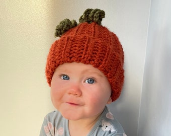 May include: A baby wearing a handmade orange crochet pumpkin hat with green stem accents. The baby is also wearing a light blue onesie with a white and pink mouse pattern. The baby is sitting on a wooden surface.