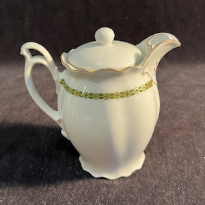 May include: White porcelain creamer with a gold and green trim. The creamer has a scalloped edge and a lid.