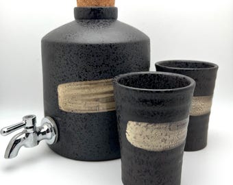May include: A black ceramic beverage dispenser with a silver spigot, a cork stopper, and two matching cups. Each piece has a textured surface and a beige horizontal band. The set is designed for serving drinks.