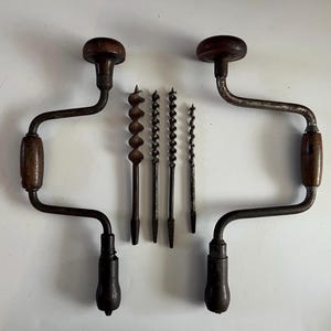 Puede incluir: Dos taladros de mano antiguos con mangos de madera y un juego de cuatro brocas. Los taladros tienen un acabado metálico oscuro con mangos y tapas de madera. Las brocas son de varios tamaños y formas.