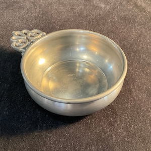 Op de afbeelding: Een zilveren tinnen kom met een decoratief handvat. De kom is rond en heeft een glad, glanzend oppervlak.