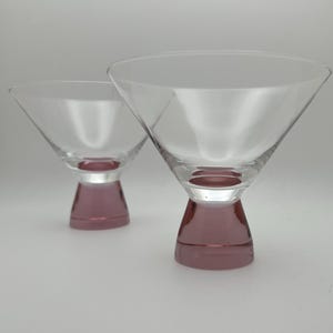 May include: Two clear martini glasses with a pink base. The glasses have a classic conical bowl shape and a short, solid pink stem. The glasses are set against a plain white background.