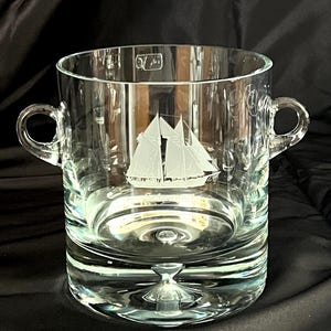 Pode incluir: Uma caneca de cerâmica branca com uma alça preta e o texto "But First, Coffee" em negrito preto. A caneca está sobre um fundo desfocado, sugerindo um ambiente acolhedor.