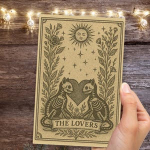 May include: A tan-colored tarot card featuring a sun with a smiling face, stars, and two cat skeletons facing each other over a heart. The card is framed by floral elements and the words "THE LOVERS" are at the bottom.
