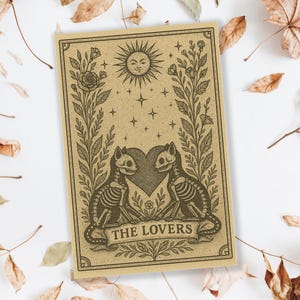 May include: A tan-colored tarot card featuring a sun with a face, stars, and two cat skeletons facing each other over a heart. The card is framed by floral elements and the words "THE LOVERS" are at the bottom.