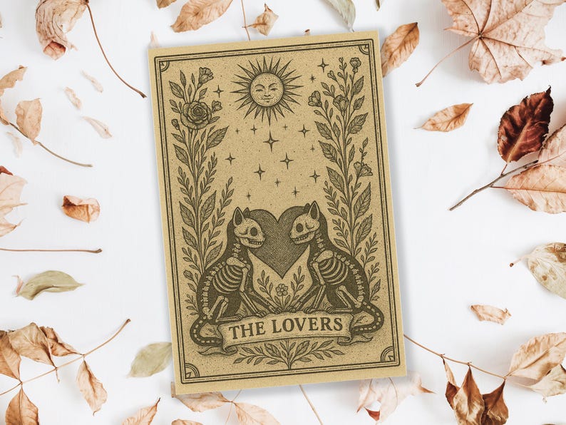 May include: A tan-colored tarot card featuring a sun with a face, stars, and two cat skeletons facing each other over a heart. The card is framed by floral elements and the words "THE LOVERS" are at the bottom.