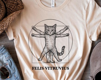 Puede incluir: Una camiseta beige con una ilustración en blanco y negro de un gato en una pose de Hombre de Vitruvio. El texto "FELIS VITRUVIUS" está impreso debajo de la imagen.