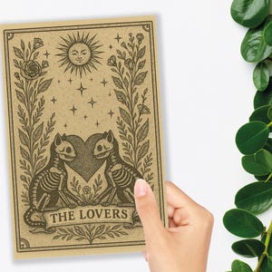 May include: A tarot card illustration featuring two skeleton cats facing each other, with a heart between them. The card has a sun, stars, and floral elements. The text "THE LOVERS" is at the bottom.