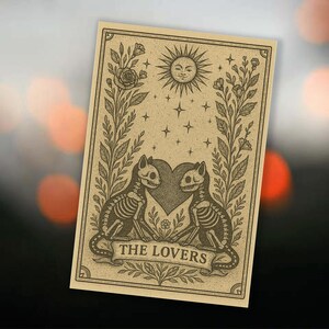 May include: A beige tarot card illustration featuring two cat skeletons facing each other, with a heart between them. Above, a smiling sun and stars are depicted. The words "THE LOVERS" are on a banner below the cats.