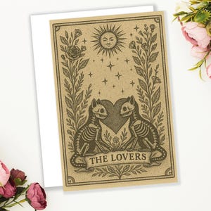 May include: A tarot card illustration featuring two skeleton cats facing each other, with a heart between them. The card has a sun, stars, and floral elements. The text "THE LOVERS" is on a banner at the bottom.