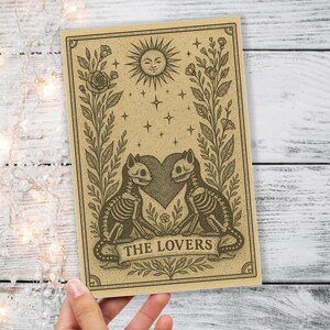 May include: A tan tarot card featuring a sun with a smiling face, stars, and floral elements. Two skeleton cats face each other, with a heart between them. The text "THE LOVERS" is at the bottom.