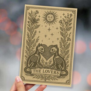 May include: A tan-colored tarot card featuring a sun with a smiling face, stars, and floral accents. Two skeleton cats face each other, with a heart between them. The banner at the bottom reads "THE LOVERS."