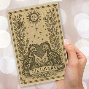 May include: A tarot card illustration featuring two skeleton cats facing each other, with a heart between them. The card has a banner that reads "THE LOVERS". The card also has a smiling sun, stars, and floral accents.