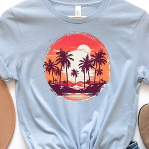 May include: Light blue t-shirt with a graphic print of palm trees and a sunset in a circle. The graphic is in shades of orange, red, and purple.