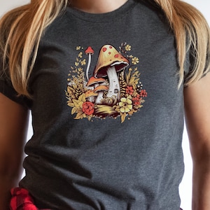 Könnte beinhalten: Ein dunkelgraues T-Shirt mit einem bunten Pilzmotiv. Die Pilze sind rot, orange und gelb mit weißen Punkten. Das Motiv ist von grünen Blättern und Blumen umgeben.
