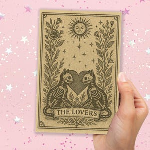 May include: A tarot card featuring a sepia-toned illustration of two skeleton cats facing each other, with a heart between them. The card has a sun with a smiling face, stars, and the text "THE LOVERS".
