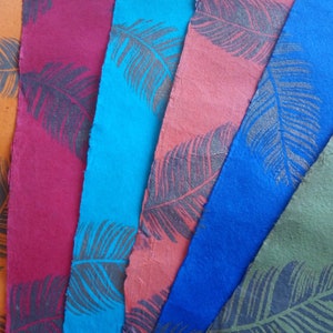 May include: A collection of six rectangular sheets of textured paper arranged in a fan pattern. Each sheet features a distinct solid color including orange, magenta, cyan, coral, blue, and olive green, all printed with a dark gray feather motif.