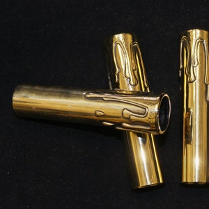 May include: A collection of five brass candle sleeves. The sleeves are cylindrical with a gold finish and a dripping wax design at the top. The candle holders are arranged on a black surface.