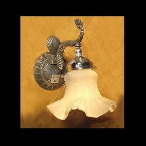 May include: An antique wall sconce with a decorative metal base and a frosted glass shade. The sconce features a detailed design with a seashell motif and a curved arm. The shade has a ruffled edge and a textured surface.