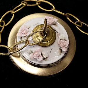 May include: A gold chain hangs from a white porcelain ceiling fixture with a floral design. The fixture has a gold metal base and a gold hook.