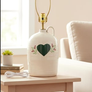 May include: Cream-colored ceramic jug lamp with a green heart design and floral accents. The lamp has a brass-colored neck and a gold-toned lamp holder. It sits on a light wood table.