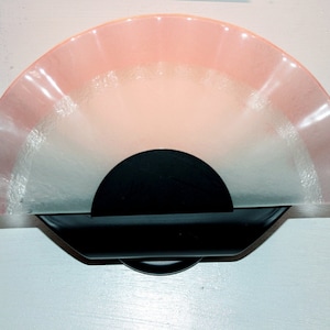 May include: A pink and white glass wall sconce with a black metal base. The glass is shaped like a fan and has a scalloped edge.