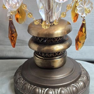 以下が含まれることがあります： ダークブロンズ調の金属製スタンドを備えたテーブルランプベース。段状の円形デザインと格子模様が特徴です。上部には、琥珀色のクリスタルティアドロップと透明な花形のガラスビーズが吊り下げられています。