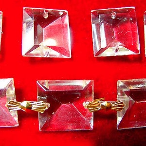 May include: A set of clear glass square crystals with gold metal connectors. The crystals are faceted and have a hole in the centre. The connectors are in the shape of a bow.