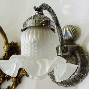 May include: A silver and white wall sconce with a frosted glass shade. The shade has a ruffled edge and a decorative design. The sconce has a decorative metal base.