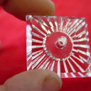 May include: A clear glass square with a raised, circular design in the center. The design has a sunburst pattern with radiating lines.